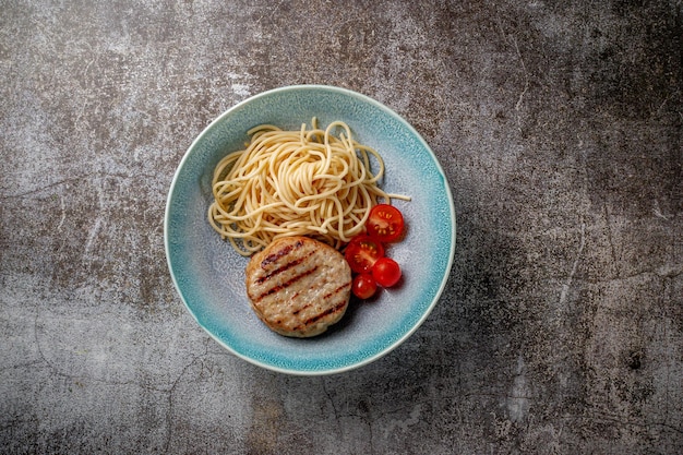 Spaghetti pasta met vleespasteitje en verse tomaten op een blauw bord tegen een grijze stenen tafel.