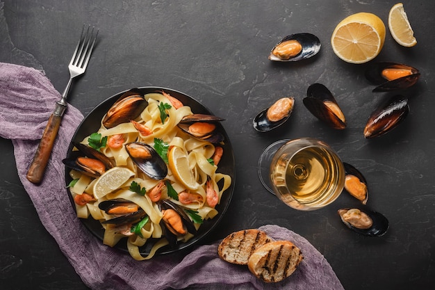 Spaghetti vongole, pasta italiana di mare con vongole e cozze, in piatto con erbe e bicchiere di vino bianco su fondo rustico in pietra.