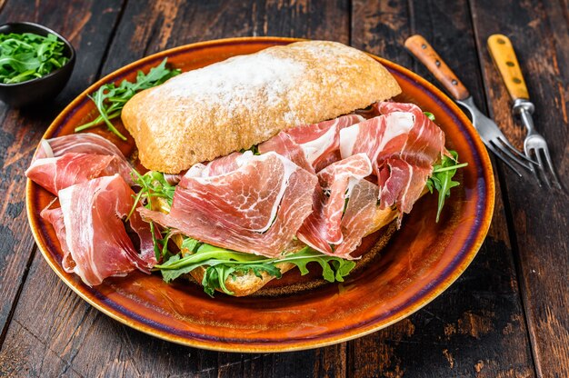 Spanish bocadillo de jamon, serrano ham sandwich on ciabatta bread with arugula. Dark wooden background. Top view.