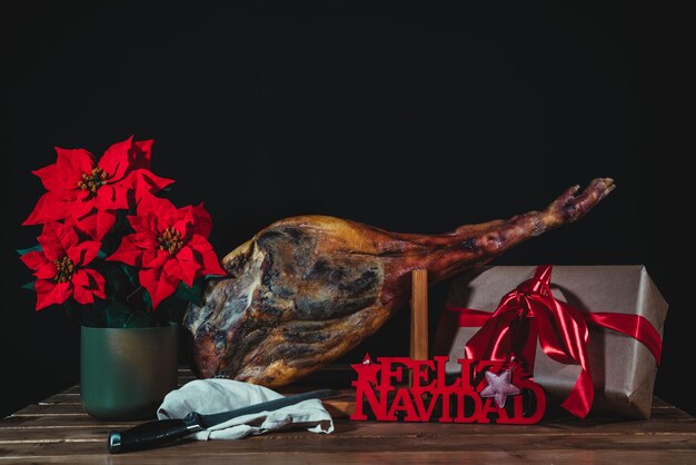 Фото Испанская ветчина с пуансеттией в подарок, знак счастливого рождества и нож на черном фоне с подарочной коробкой