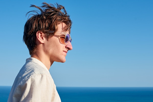 Spanish man with long hair, glasses and a beige shirt glaring to the side on ocean and sky