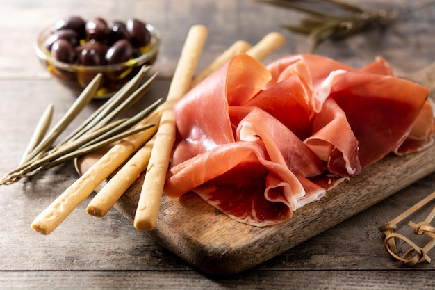 Spanish serrano ham with olives and breadstick on wooden table background