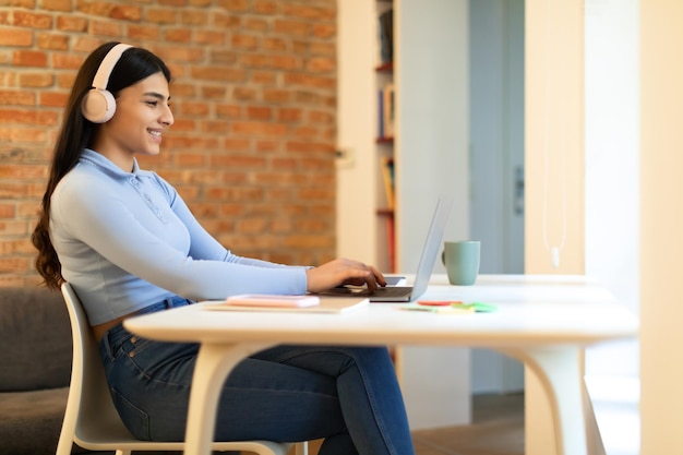 Spanish teenager lady learning with laptop computer wearing headphones doing homework while sitting