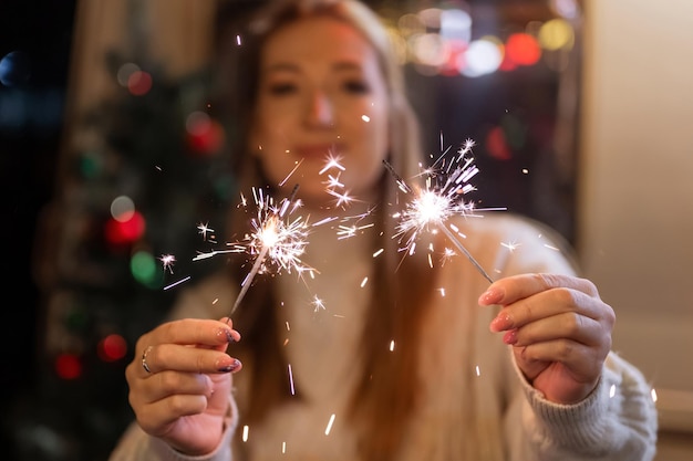 新年とクリスマスイブの夜に屋外のキャンピングトレーラーの家の背景にニットウールの白い居心地の良いセーターで女性を保持している女性の手でスパークラーベンガル火棒。きらめく火花