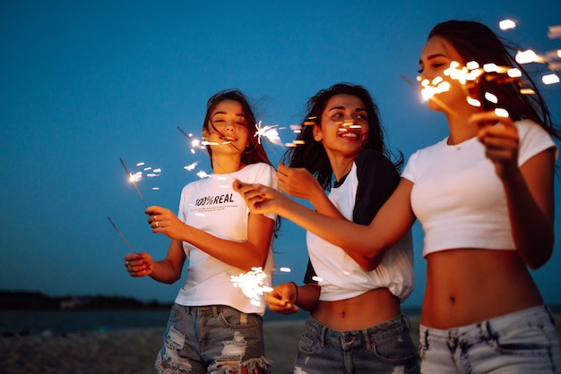 ビーチで若い女の子の手に花火。花火とビーチでパーティーを楽しんでいる3人の女の子。夏の休日、休暇、リラックス、ライフスタイルのコンセプト。