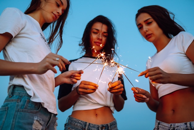 ビーチで若い女の子の手に花火。花火とビーチでパーティーを楽しんでいる3人の女の子。夏の休日、休暇、リラックス、ライフスタイルのコンセプト。