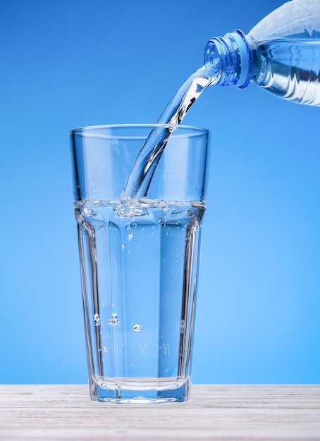 L'acqua frizzante si versa dalla bottiglia nel bicchiere grande. sfondo blu, tavolo bianco.