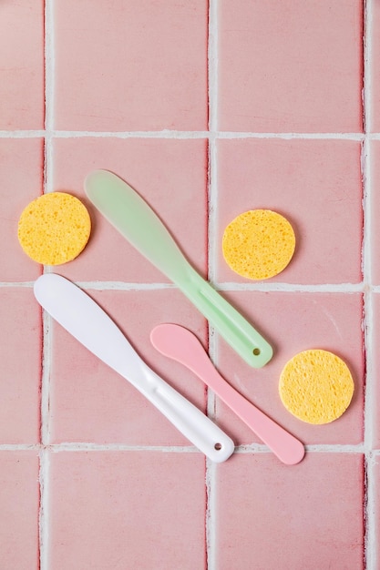 Spatulas for applying face mask or cream and sponges for washing face on pink tiled background top view Tools for cosmetology