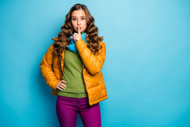 Don't speak! photo of pretty curly lady holding finger on lips
asking keep secret in privacy wear casual yellow overcoat scarf
violet trousers isolated blue color wall