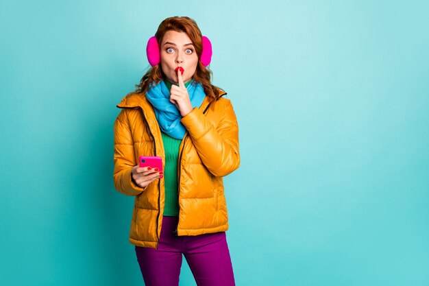 Don\'t speak! portrait of funny lady hold telephone finger on\
lips tell secret information ask to keep silence wear ear muffs\
yellow overcoat scarf pants.