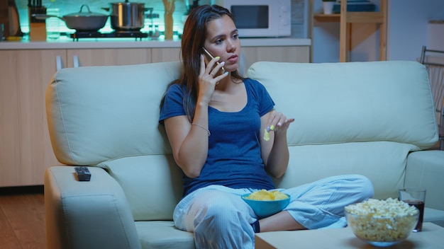 Speaking on phone holding snacks bowl and eating snacks in front of TV. Happy, relaxed, lonely lady in pajamas enjoying the evening sitting on comfortable couch in front of television using smartphone