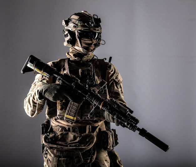 special forces fighter in battle uniform and helmet with radio headset, face mask and ballistic glasses, standing with equipped laser sight and silencer service rifle studio portrait isolated on white