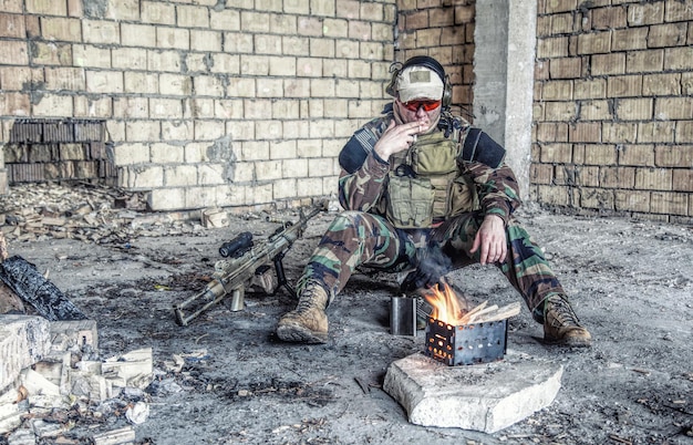 Special forces-soldaat na het gevecht zittend bij het vuur in een verwoest gebouw rokende sigaret