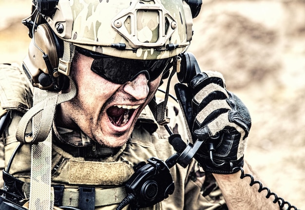 Soldato delle forze speciali, operatore di comunicazioni militari o manutentore in casco e occhiali, urlando in radio durante la battaglia nel deserto. richiamare i rinforzi, riferire la situazione sul campo di battaglia