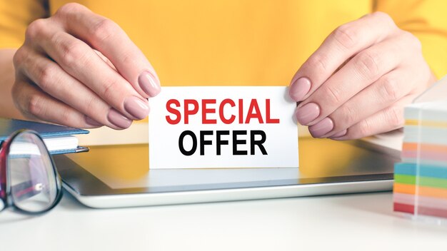Special offer is written on a white business card in a woman's hands. Glasses, tablet and block with multi-colored paper for notes.