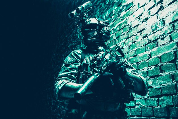 Солдат сил специальных операций, боец штурмовой группы по борьбе с терроризмом, с прибором ночного видения на шлеме и служебной винтовкой, сдержанный выстрел в помещении, кирпичная стена