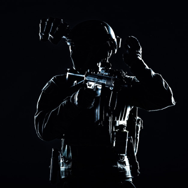 Special operations soldier, SWAT team fighter in mask and glasses, equipped night vision device, armed short barrel service rifle, looking back and showing freeze hand signal, low key studio shoot