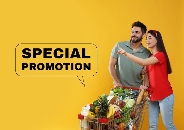 黄色の背景に食料品がいっぱい入ったショッピングカートを持つ特別なプロモーションの若いカップル
