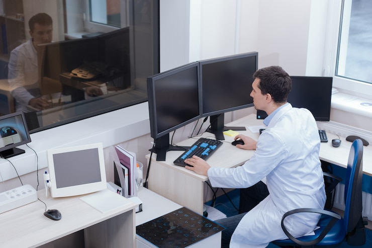 Photo special software. serious nice handsome doctor sitting at the table and working on the computer while controlling the process of ct scanning