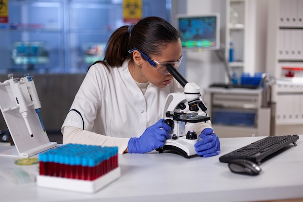 Specialist biochemist woman analyzing blood sample under microscope developing vaccine against coronavirus in microbiology hospital laboratory. Chemist doctor researcher analyzing virus expertise