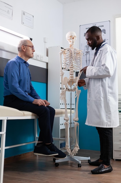 Specialist chiropractor doctor showing human skeleton explaining back pain to old man patient discussing sickness symptoms during medical appointment in hospital office. Osteoporosis treatment