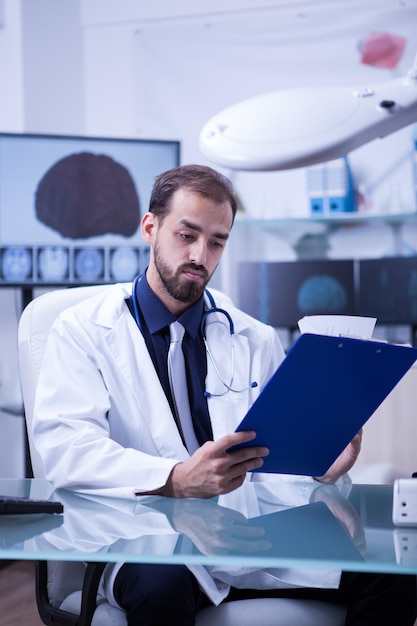 Specialist doctor checking his clipboard sitting at his desk in the hospital cabinet. Monitor with a 3d brain.