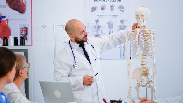 Specialist doctor in white coat showing to colleagues body bone functionson using skeleton model. Medic pointing and explaining to coworkers medical procedures sitting in hospital meeting room.