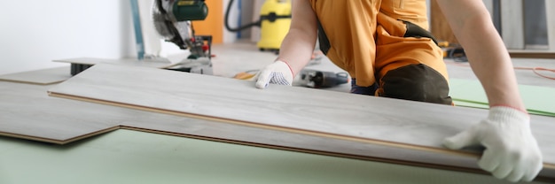 Specialist hold laminate in his handand put it on floor. Man in overalls and helmet make repairs in room.