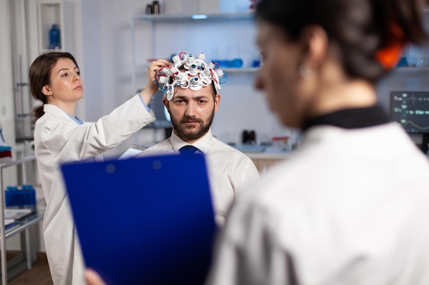 연구원 여성이 eeg 헤드셋을 조정하는 동안 남성 환자에게 신경 치료를 설명하는 클립보드를 들고 있는 전문 신경과 전문의입니다. 뇌 활동 진화를 모니터링하는 의사