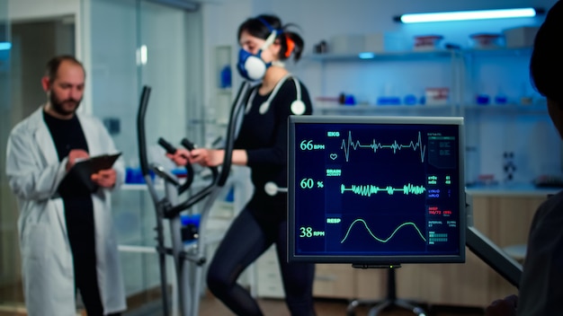 Photo specialist sport researcher monitoring heart rate on athlete while woman with mask running on cross trainer talking with medical doctor