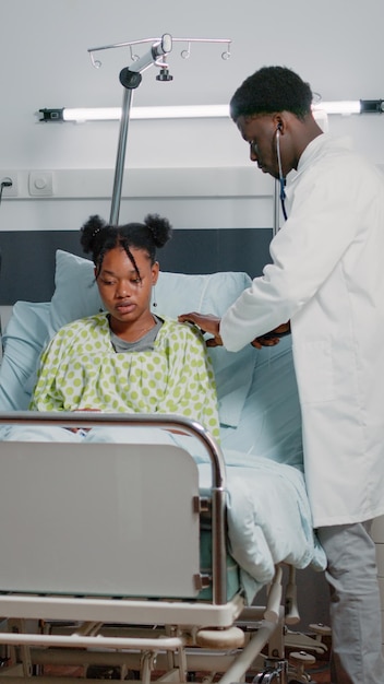 Specialist using stethoscope to examine heartbeat and pulse of patient with disease. General practitioner checking up on diagnosis of young adult in bed while medical assistant helping