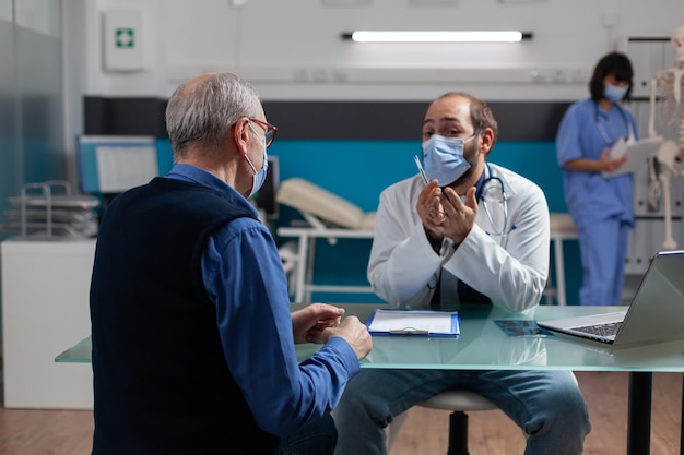 Specialist with face mask consulting elder person in medical cabinet during pandemic. Professional medic explaining health care diagnosis to mature patient at checkup appointment.