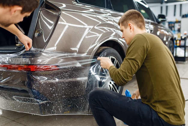 전문가들은 리어 범퍼에 자동차 보호 필름을 적용합니다. 자동차 페인트를 스크래치로부터 보호하는 코팅 설치. 차고의 새 차량, 튜닝
