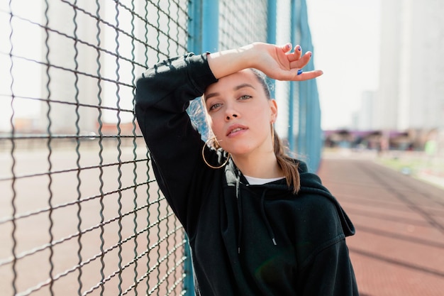 Spectacular charming lady in black hoodie is posing at camera outdoor in sunlight She is touching her head and smiling