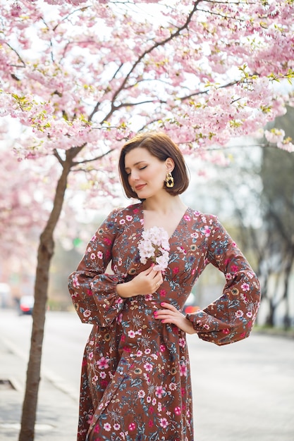 桜を背景に明るいドレスを着た見事な女性が立っています。美しい衣装を着た黒髪の女性が歩きながら通りに微笑む