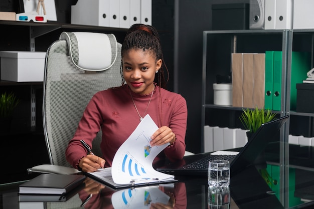 Spectacular young black businesswoman signs documents at table in office