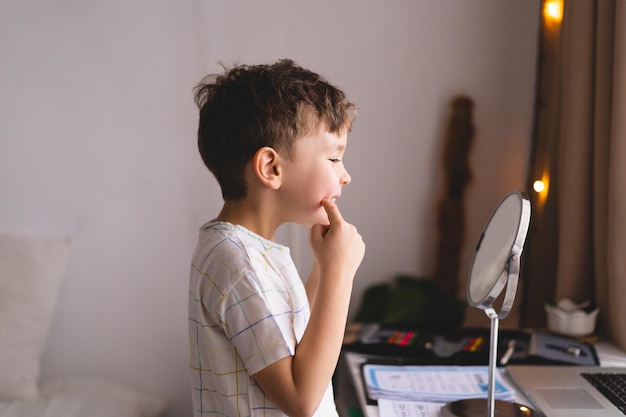 言語療法の概念音を発音するかわいい男の子Oラップトップを介してオンライン言語療法のレッスンをしている鏡を見ている