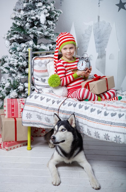 Speels glimlachend gelukkig schattig meisje gekleed in gestreepte pyjama's met cadeau, klok en hond zittend in ingerichte nieuwjaarskamer thuis. Kerst goed humeur. Lifestyle, familie en vakantie 2018 concept