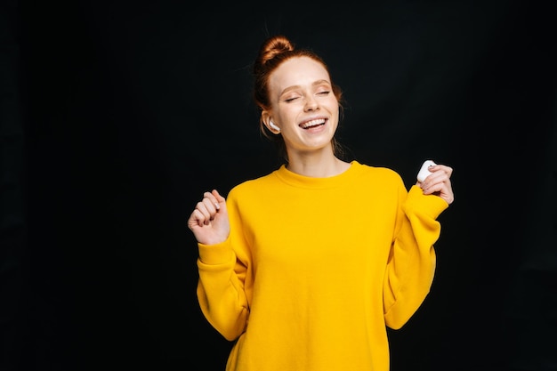 Speelse jonge vrouw met draadloze koptelefoon die danst terwijl ze naar populaire liedjes luistert met gesloten ogen