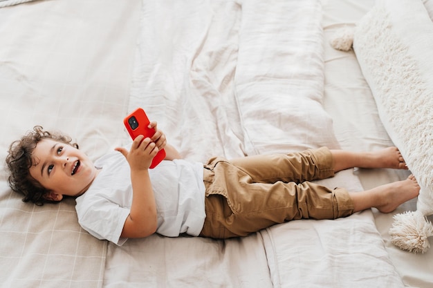 Speelse krullende Braziliaanse jongen in wit t-shirt en broek liggend op bed lachend met telefoon vasthoudend met lege ruimte voor advertentie Dromende kaukasische knappe jongen die mobiel thuis gebruikt Jeugd en gadgets
