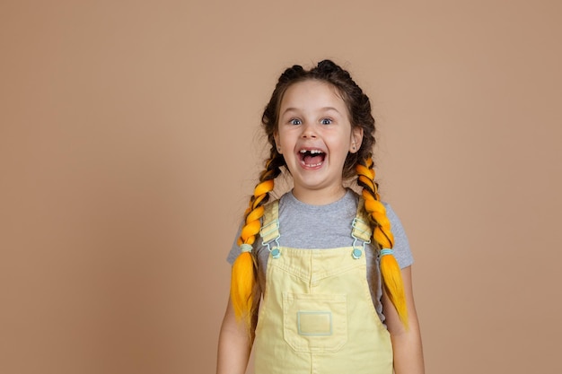 Speelse opgewonden kleine vrouw met gele kanekalon staartjes glimlachend met geopende mond met ontbrekende tand dragen gele jumpsuit en grijze tshirt op beige achtergrond