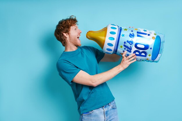 Speelse roodharige man die een flesvormige ballon vasthoudt terwijl hij tegen een blauwe achtergrond staat