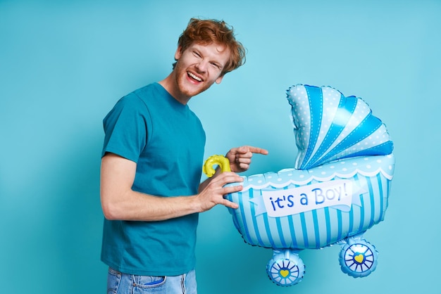 Speelse roodharige man met een babywagentje in de vorm van een ballon terwijl hij tegen een blauwe achtergrond staat