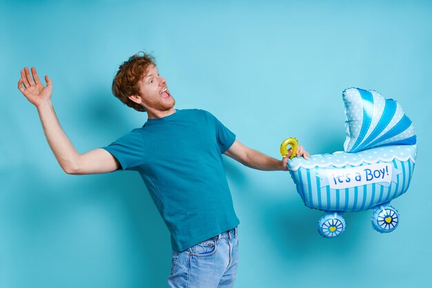 Speelse roodharige man met een babywagentje in de vorm van een ballon terwijl hij tegen een blauwe achtergrond staat