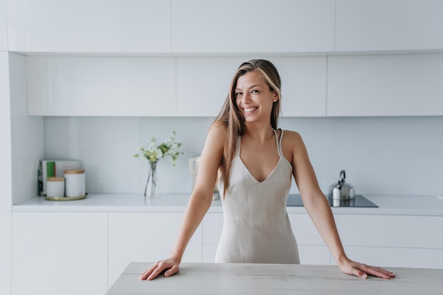 Speelse sexy blanke vrouw in nachtjapon staande in de keuken leunt op tafel kijkt naar de camera glimlacht