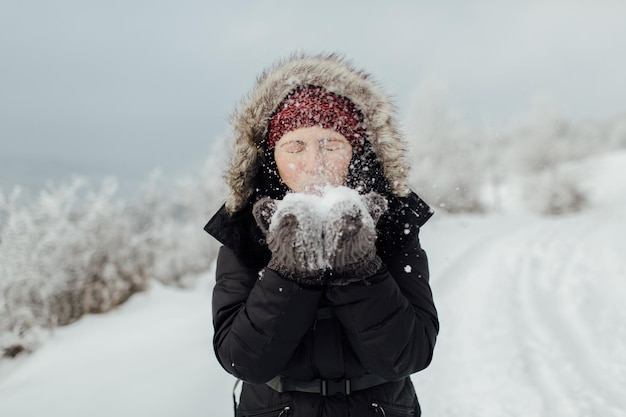 Speelse vrouw blaast sneeuw uit haar handen terwijl ze op de berg staat