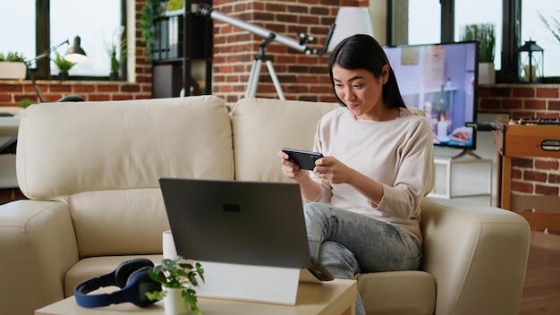 Speelse vrouw die thuis op de bank zit tijdens het spelen van arcadespellen op smartphone. Competitief ogende jonge volwassen persoon die geniet van mobiel gamen terwijl hij ontspant in de woonkamer op de bank.