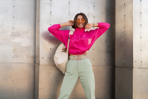 Speelse vrouw met plezier in neon roze jasje over grijze metalen stedelijke muur