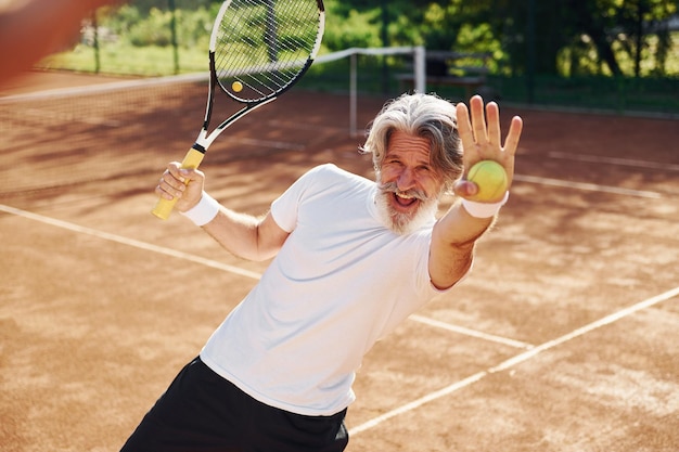Speelspel Senior moderne stijlvolle man met racket buitenshuis op tennisbaan overdag