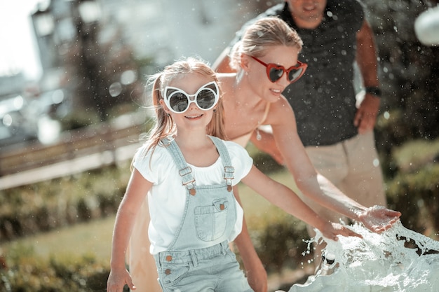 Speeltijd met ouders. glimlachend meisje spelen met fontein spatten staande voor haar ouders op de hete zomerdag.